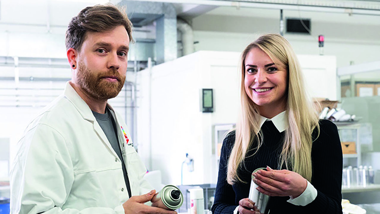 Zwei Personen lächeln in die Kamera und halten Spraydosen in den Händen. 

