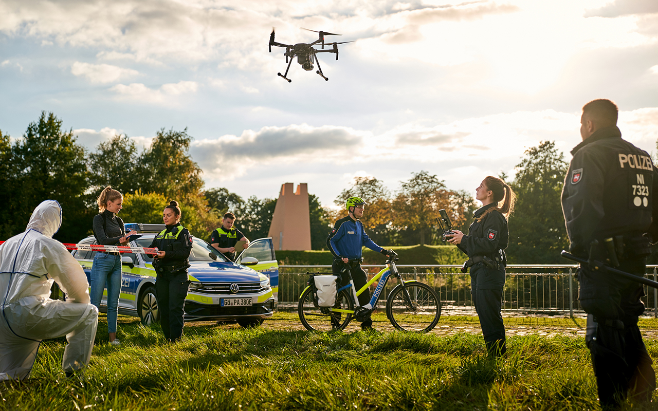 Außenaufnahme: Mehrer Polizeibeamte bei Ihrer Arbeit, am Himmel schwebt eine Drohne, ein Polizeiwagen ist zu sehen, jemand von der Spurensicherung im Schutzanzug und zum Beispiel ein Fahrradpolizist.