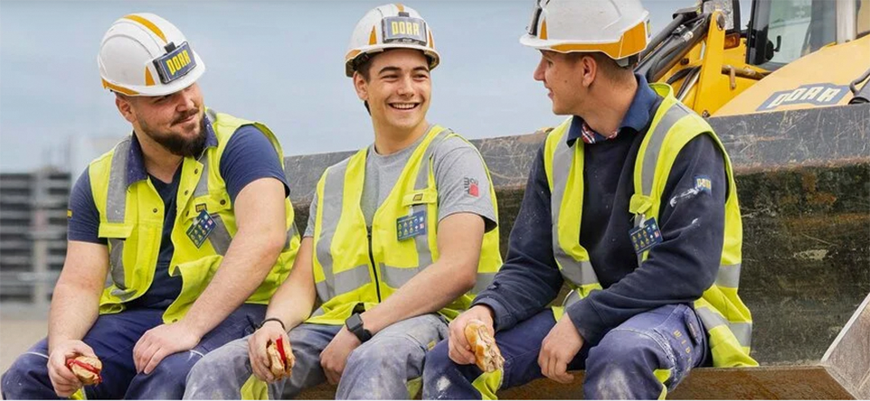 Drei Mitarbeitende von PORR in Arbeitskleidung in einer Baggerschaufel auf einer Baustelle sitzend
