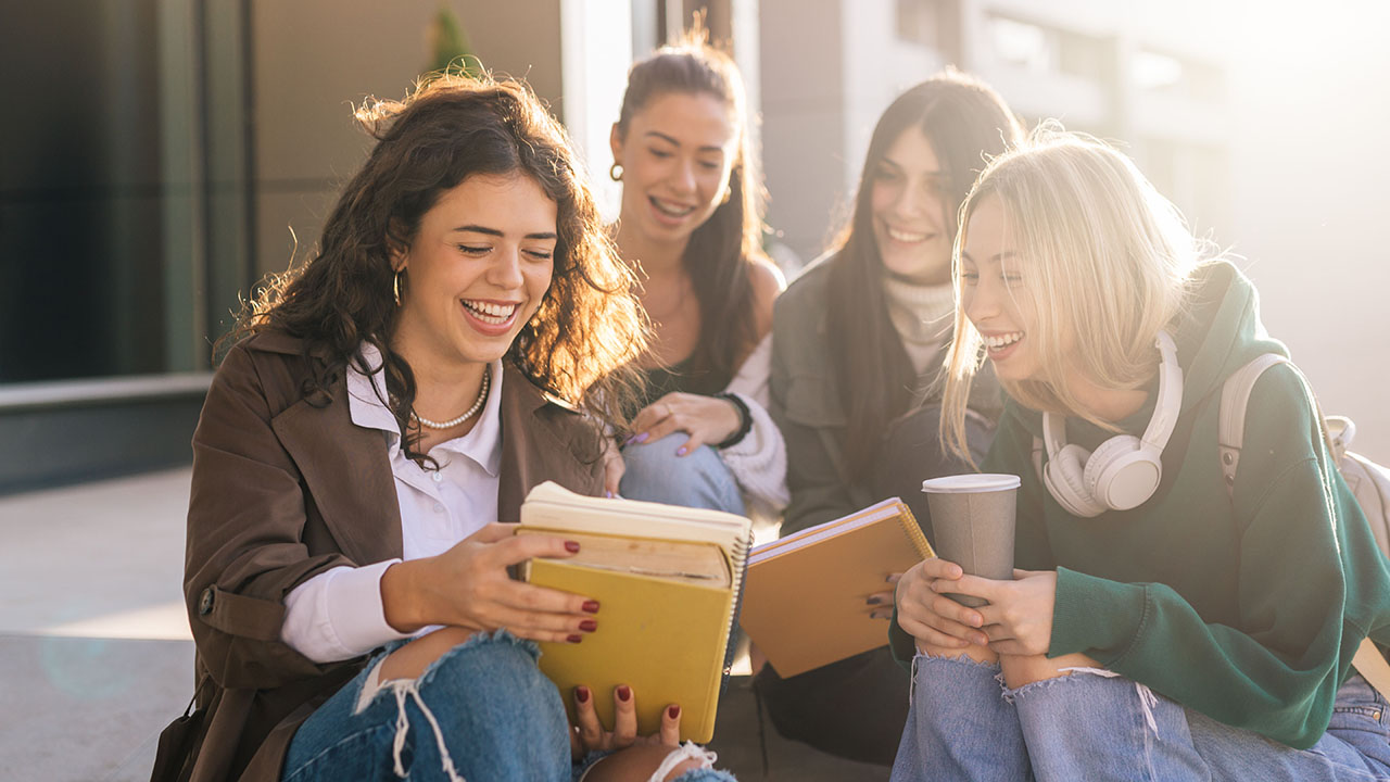Gruppe von lachenden jungen Menschen sitzt auf einer Treppe und guckt sich Unterlagen an