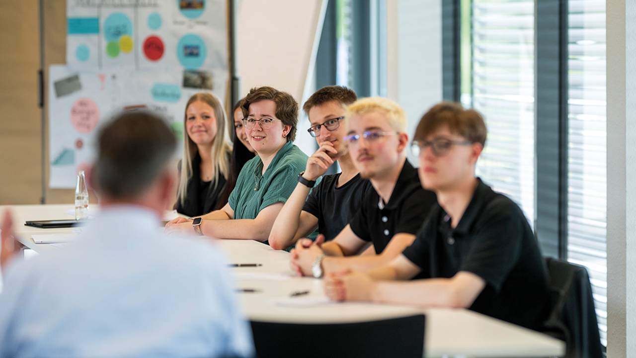 Mehrere junge Menschen sitzen bei einer Besprechung mit einem Ausbilder an einem großen Tisch.