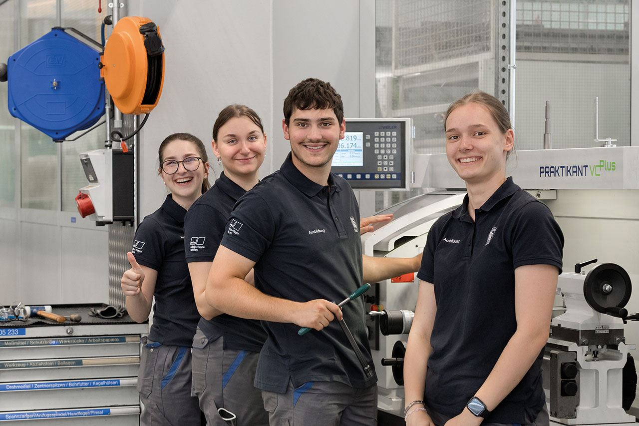 Das Bild zeigt vier Personen in Arbeitskleidung, die vor einer Maschinensteuerungsmöglichkeit in einer Werkstatt stehen. Sie blicken alle lächelnd in die Kamera.