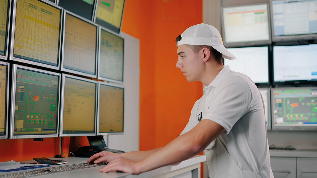 Junge Person in weißem T-Shirt und weißem Basecap steht vor mehrerem Monitoren.
