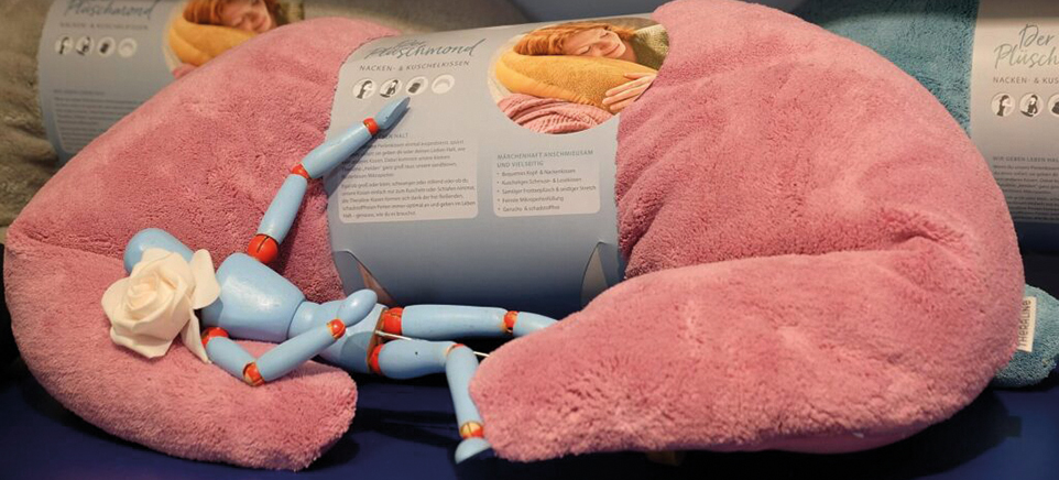 Ein großes, halbmondförmiges, rosafarbenes Kuschelkissen, das offenbar als Nacken- und Kuschelkissen dient. Vor dem Kissen liegt eine kleine, hellblaue Holz- oder Gliederpuppe mit roten Gelenken. Diese Puppe trägt eine weiße Rose als Kopfschmuck.