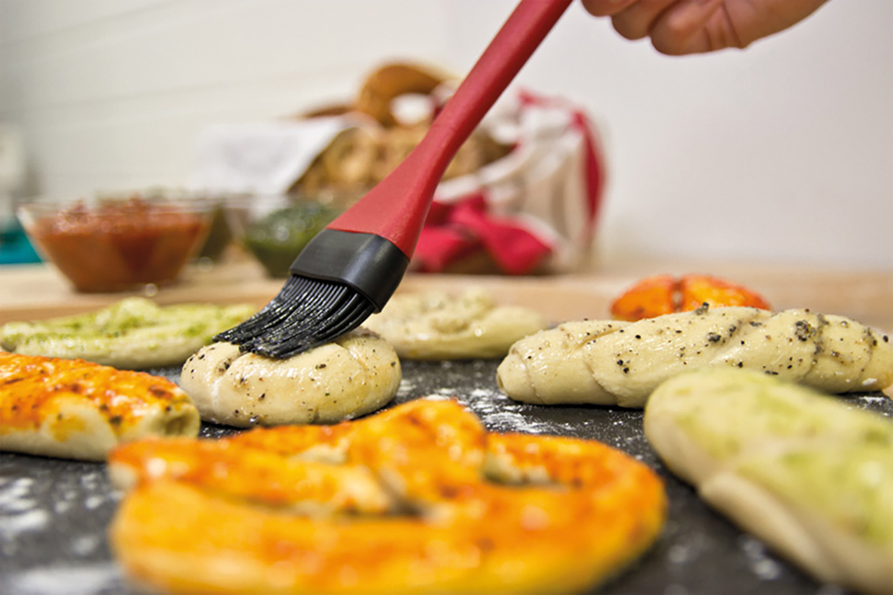 Verschiedene Kleingebäcke werden mit einem roten Backpinsle mit einer Kräutermischung eingestrichen.