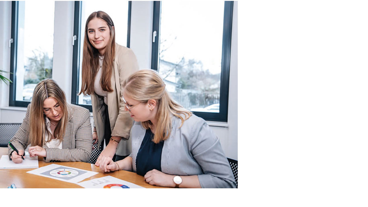 Drei Personen sitzen bzw. stehen an einem Schreibtisch und arbeiten an Diagrammen.