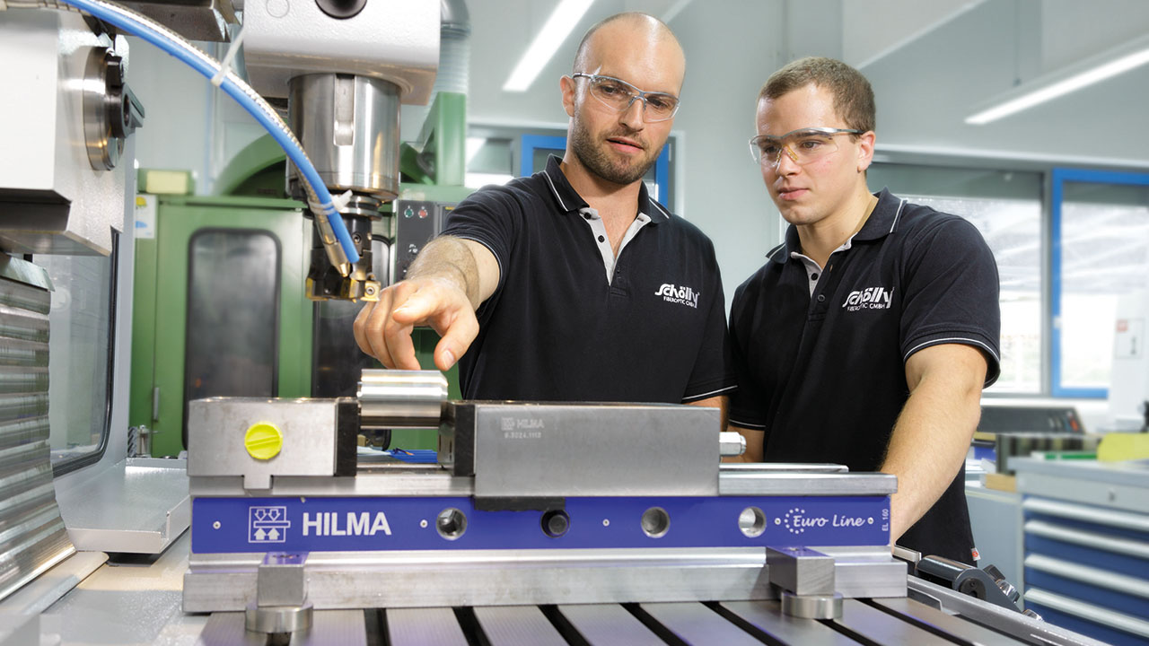 Zwei junge Menschen in Poloshirts mit Firmenlogo sehen sich eine Maschine in der Produktionsstätte an