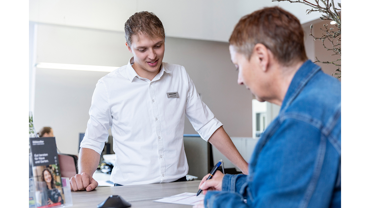 Zwei Menschen an einer Theke. Der Mitarbeiter trägt ein Hemd. Der Kunde schreibt etwas auf einem Papier