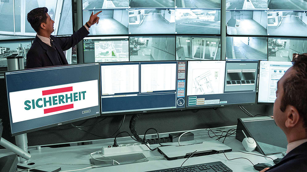 Eine Person sitzt in einem Kontrollraum mit vielen Überwachungsmonitoren. Eine weitere Person zeigt auf einen der Monitore.