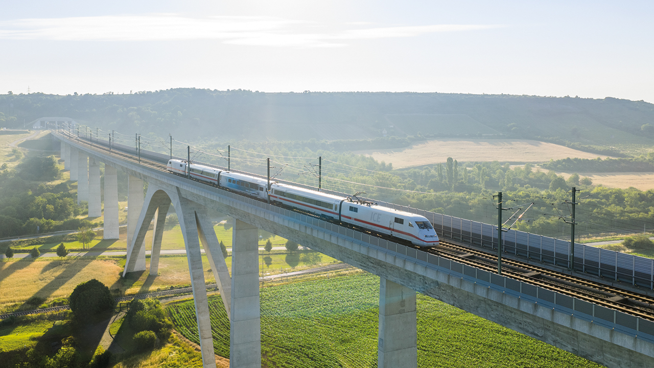 Foto eines Zuges der gerade in einer sommerlichen Landschaft über eine Brücke fährt.