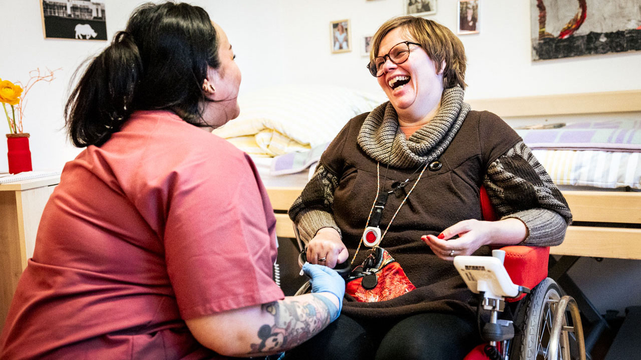 Sitzende Person mit langen Haaren und Tattoos im Gespräch mit einer lachenden Person im Rollstuhl, im Hintergrund ein Pflegebett