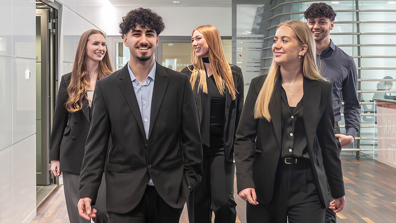 Fünf junge Mitarbeitende der Sparkasse Hannover laufen versetzt hintereinander vorn ein junger Mann neben einer Frau dahinter zwei junge Frauen und ein junger Mann auf einem Gang in der Sparkasse Hannover.
