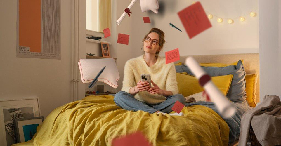 Frau sitzt verträumt auf den Bett. Um sie herum schweben Schreibtischgegenstände.