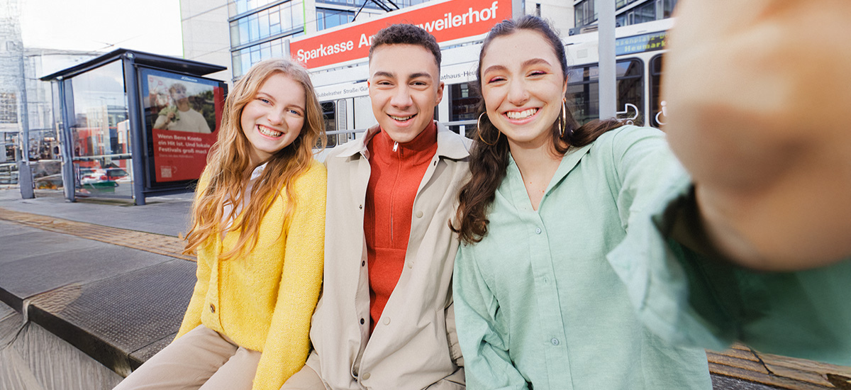 Drei junge Menschen sitzen auf einer Bank vor der Bankfiliale und lächeln in die Kamera