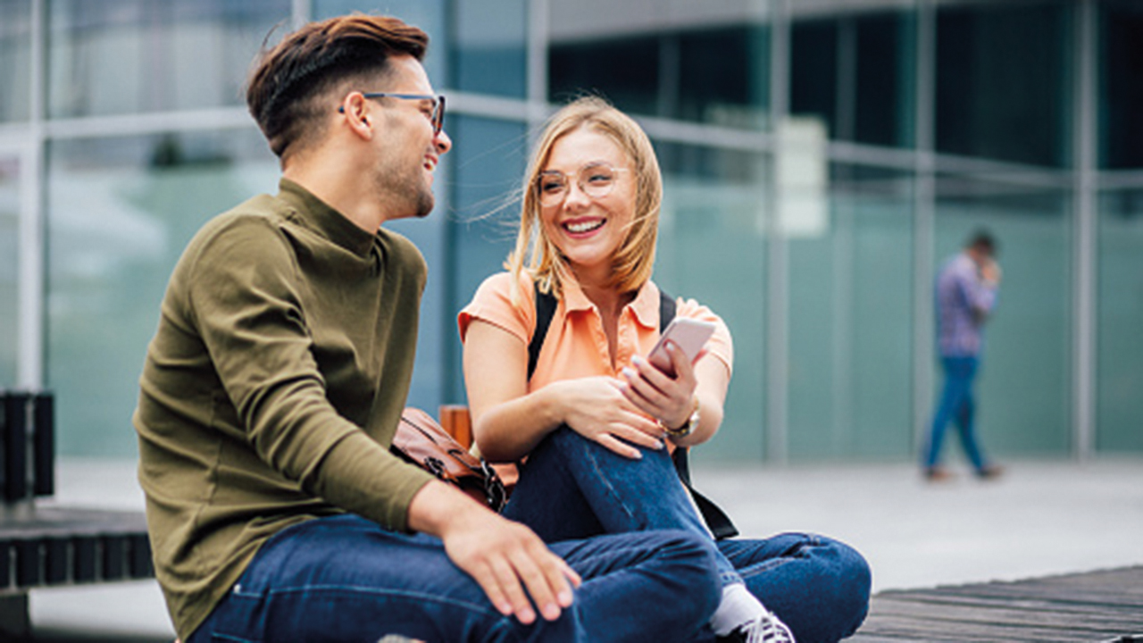 Zwei Studierende sitzen vor einem Unigebäude und sind in ein freundliches Gespräch vertieft.
