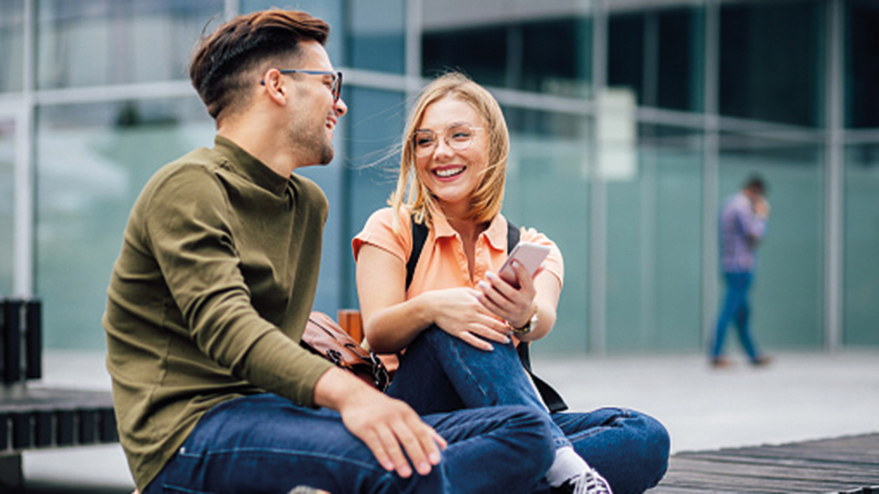 Zwei Studierende sitzen vor einem Unigebäude und sind in ein freundliches Gespräch vertieft.