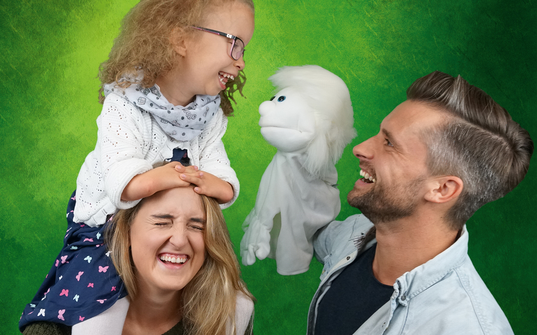 Eine junge Frau auf deren Schultern ein kleines Mädchen sitzt. Beide lachen herzlich. Daneben ein junger Mann mit einer weißen Handpuppe der das Mädchen anlächelt.