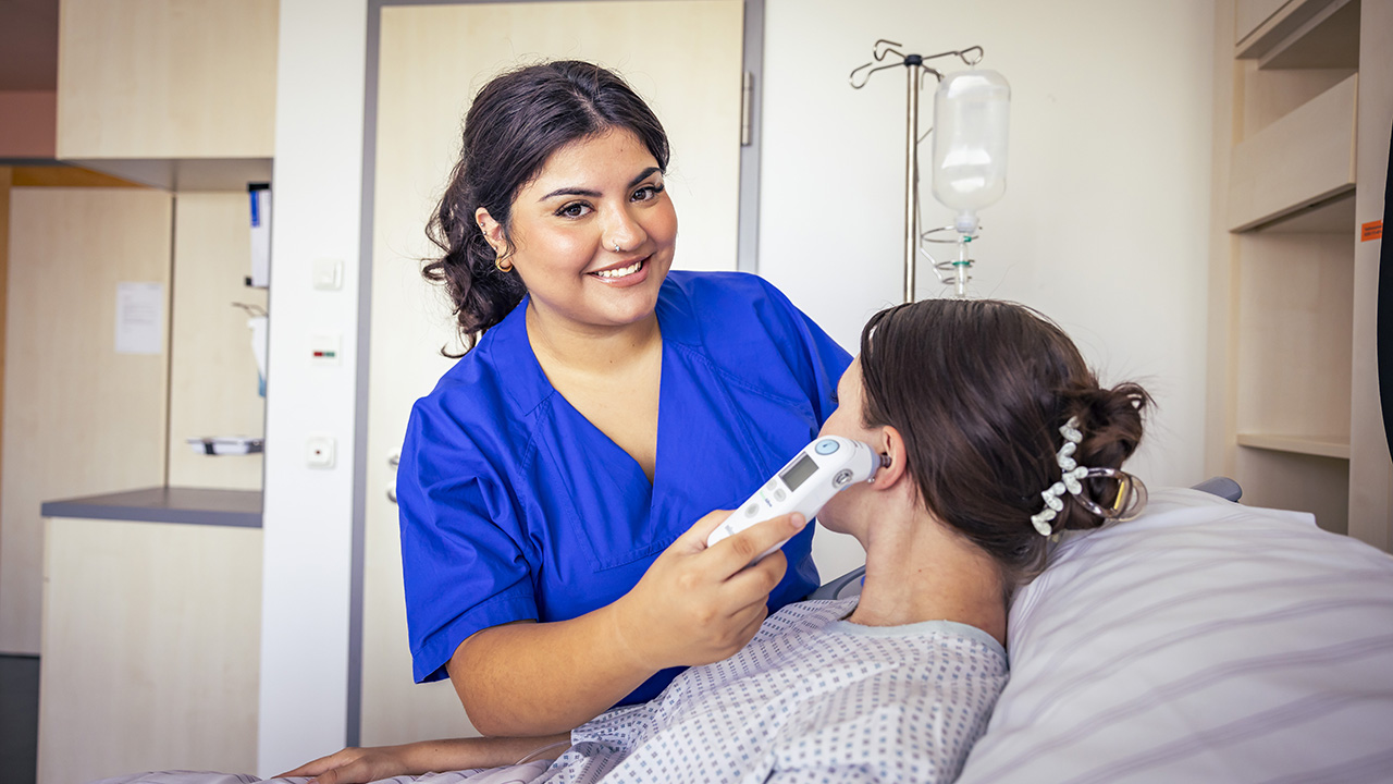 Eine Pflegeperson misst bei einer Patientin die Temperatur mit einem Fieberthermometer am Ohr.