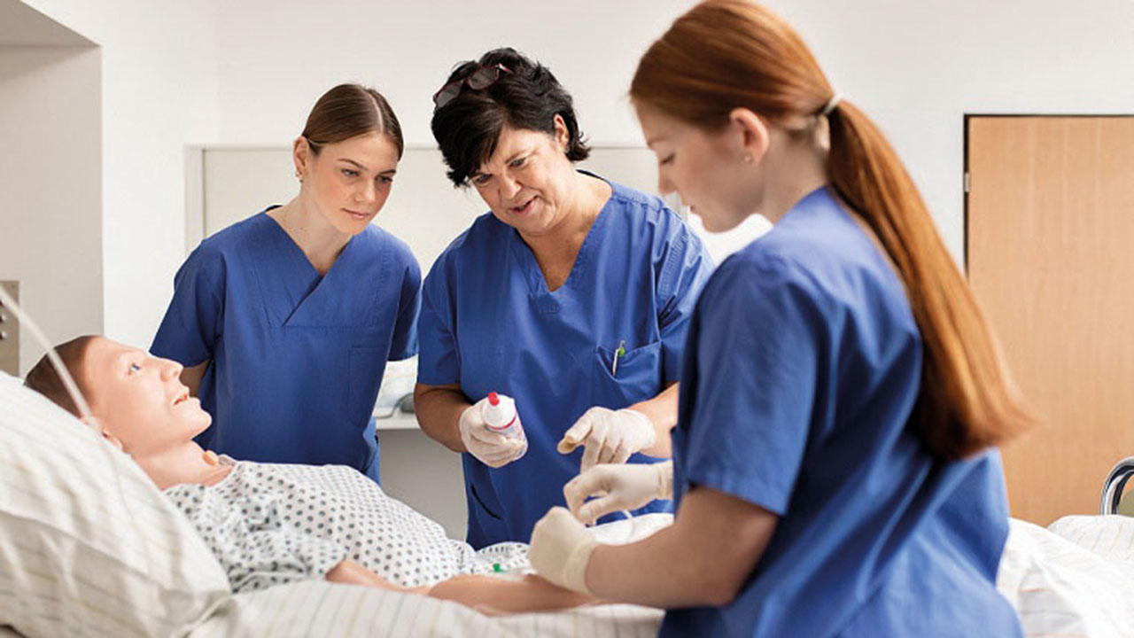 Drei Menschen in blauer Pflegebekleidung am Krankenbett, Auszubildende übt die Versorgung an einer Pflegepuppe