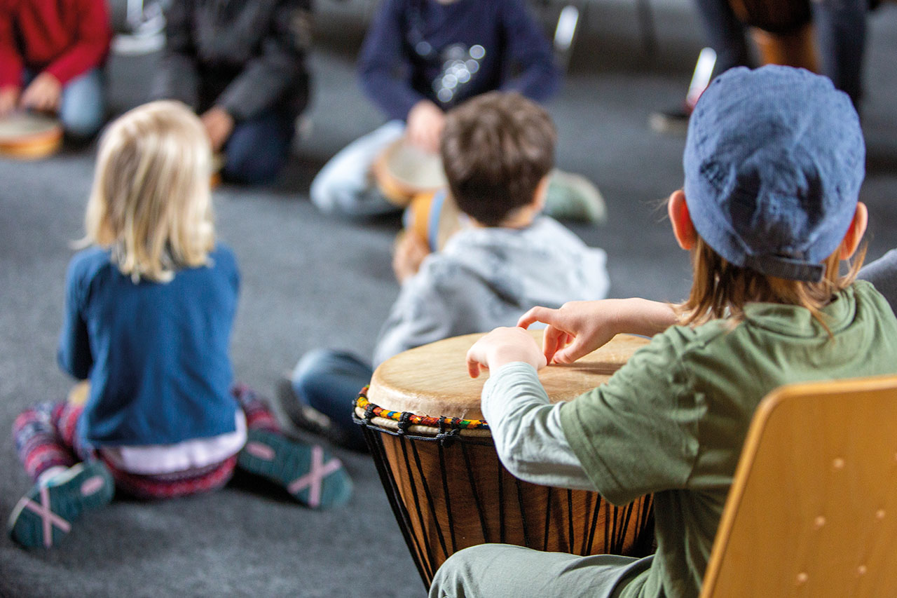 Kinder sitzen auf einem Fußboden. Ein Kind sitzt auf einem Stuhl und hat eine Trommel vor sich.