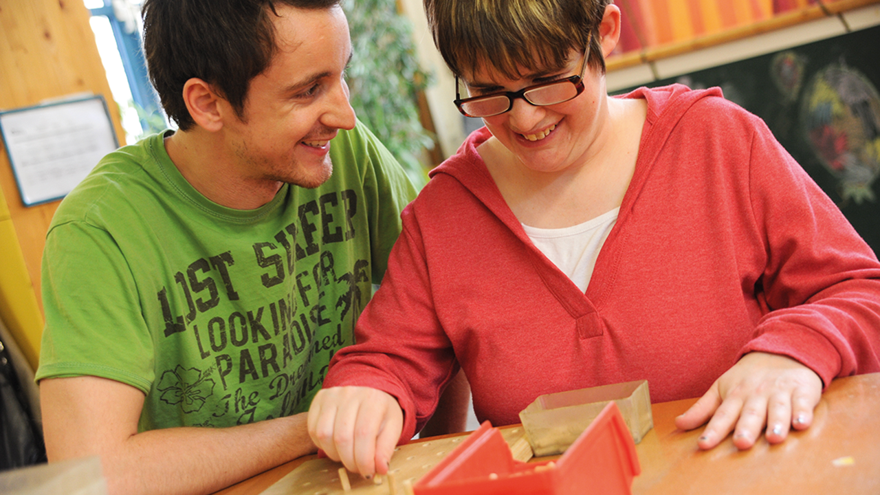 Betreuer unterstützt einen Schützling bei einer feinmotorischen Übung