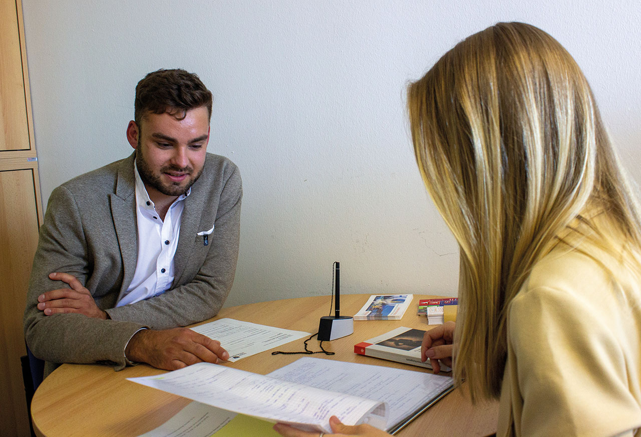 Zwei Personen sitzen an einem Schreibtisch in einem Büro. Eine Person blickt in eine Mappe. Auf dem Tisch liegen Flyer.