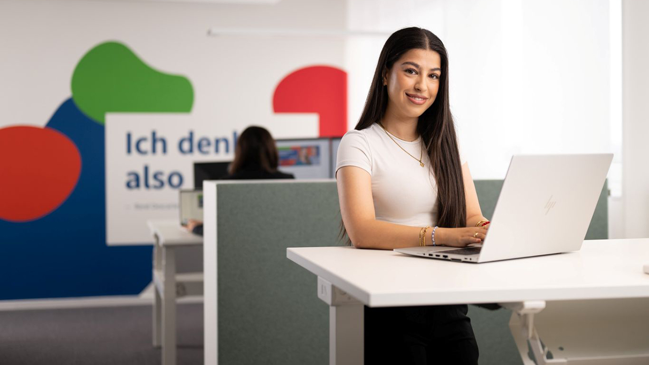 Frau steht an einem Schreibtisch mit einem Laptop. Es ist ein Büroraum mit einer bunten Wand im Hintergrund.