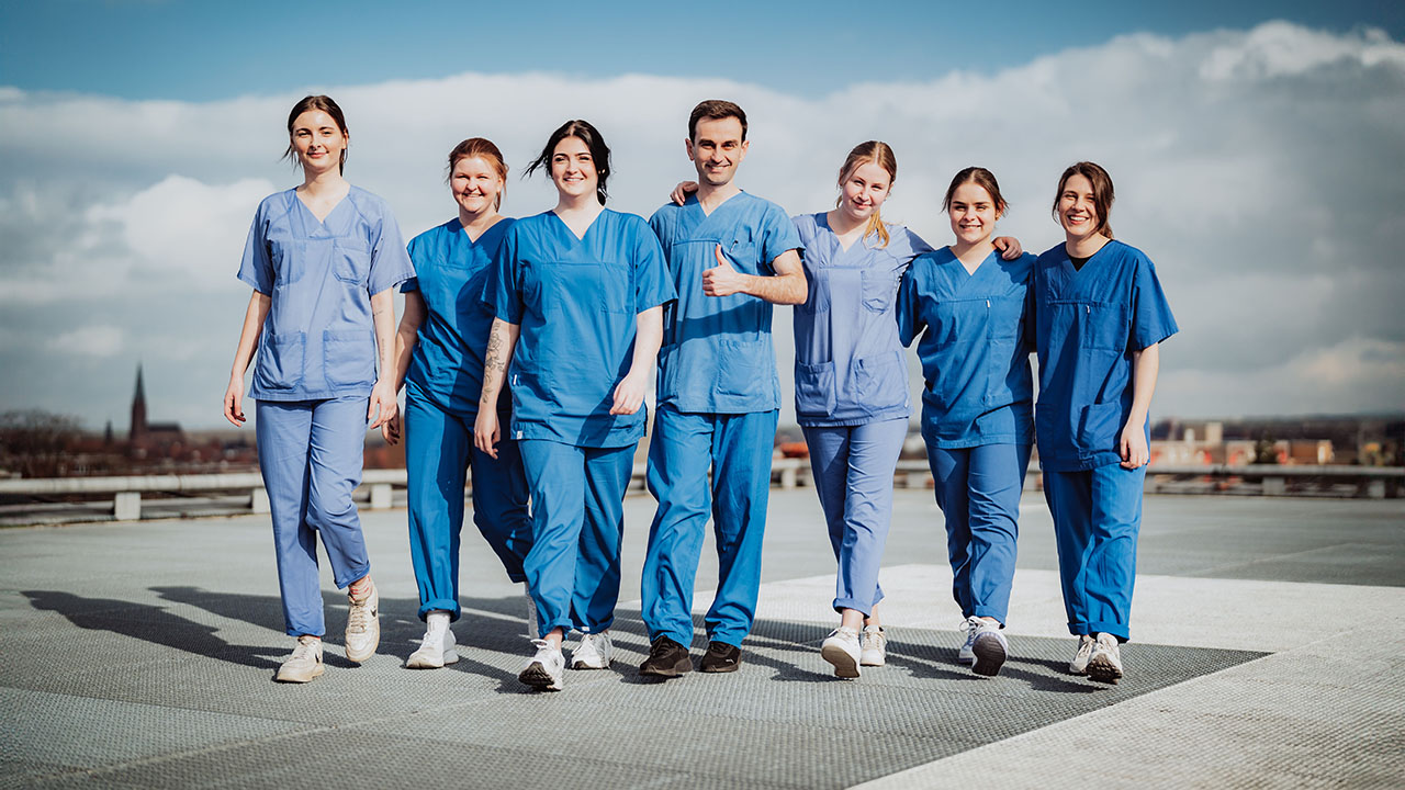 Sieben junge lächelnde Personen in blauer Pflegebekleidung auf dem Dach des Klinikums