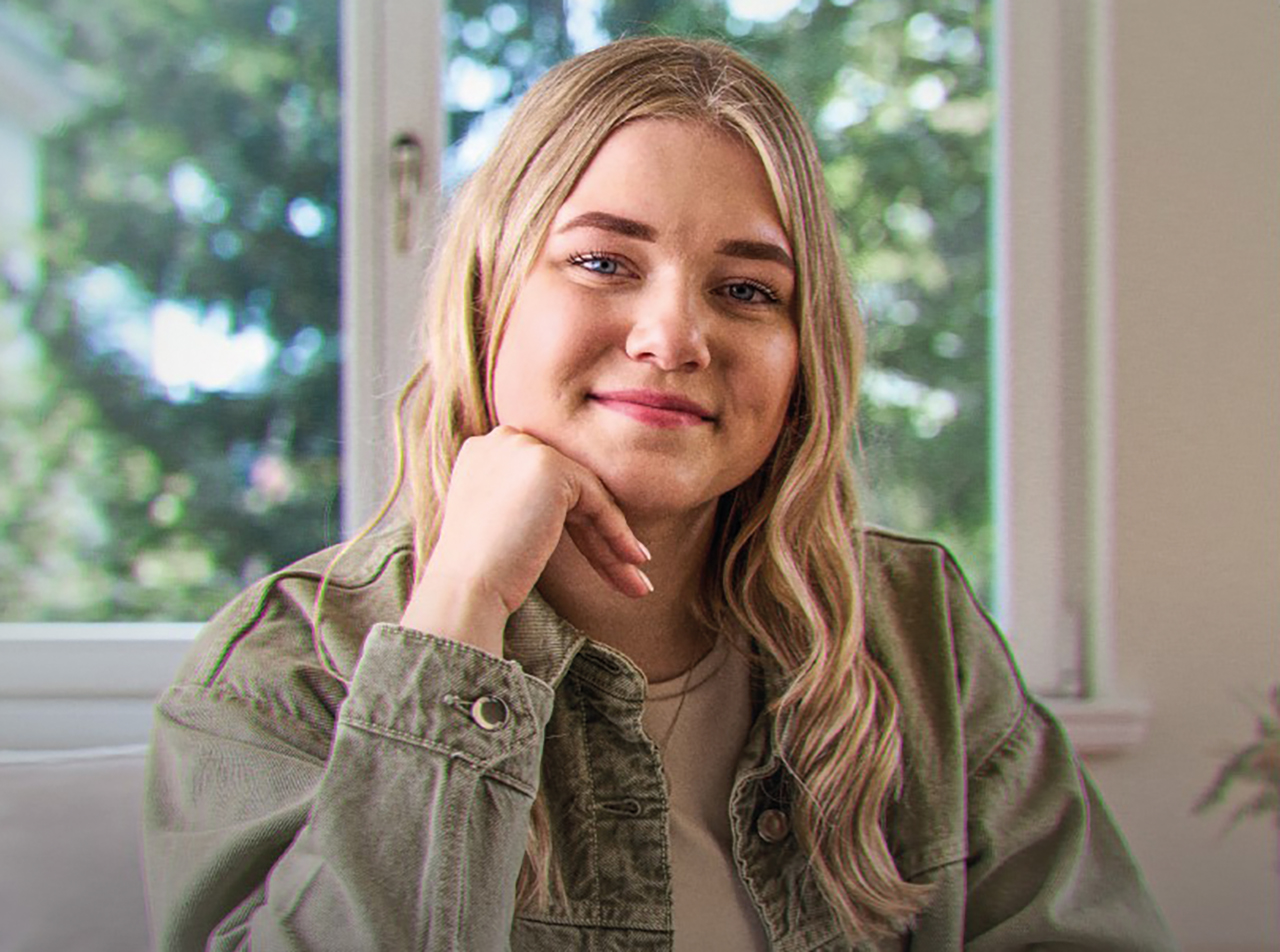 Junges Mädchen mit blonden Haar sitzt vor einem Fester und stützt sich Kopf leicht auf ihre Hand.