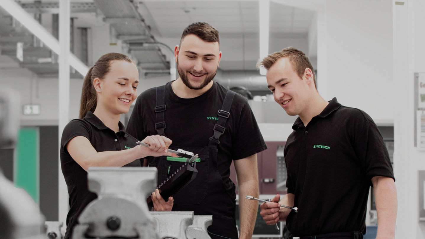 Drei Personen in Arbeitskleidung stehen in einer Werkstatt und betrachten gemeinsam ein Werkzeugteil.