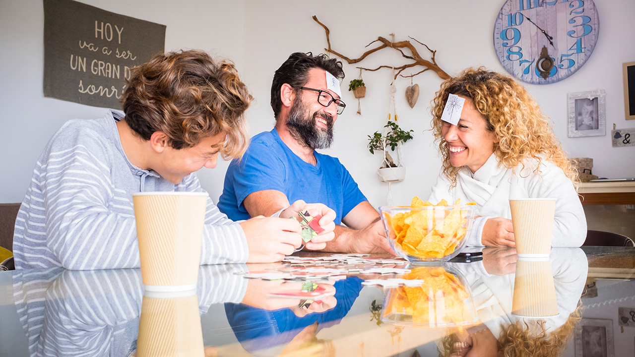 Drei Menschen sitzen am Tisch auf dem Snacks stehen. Sie spielen ein Kartenspiel und lachen.