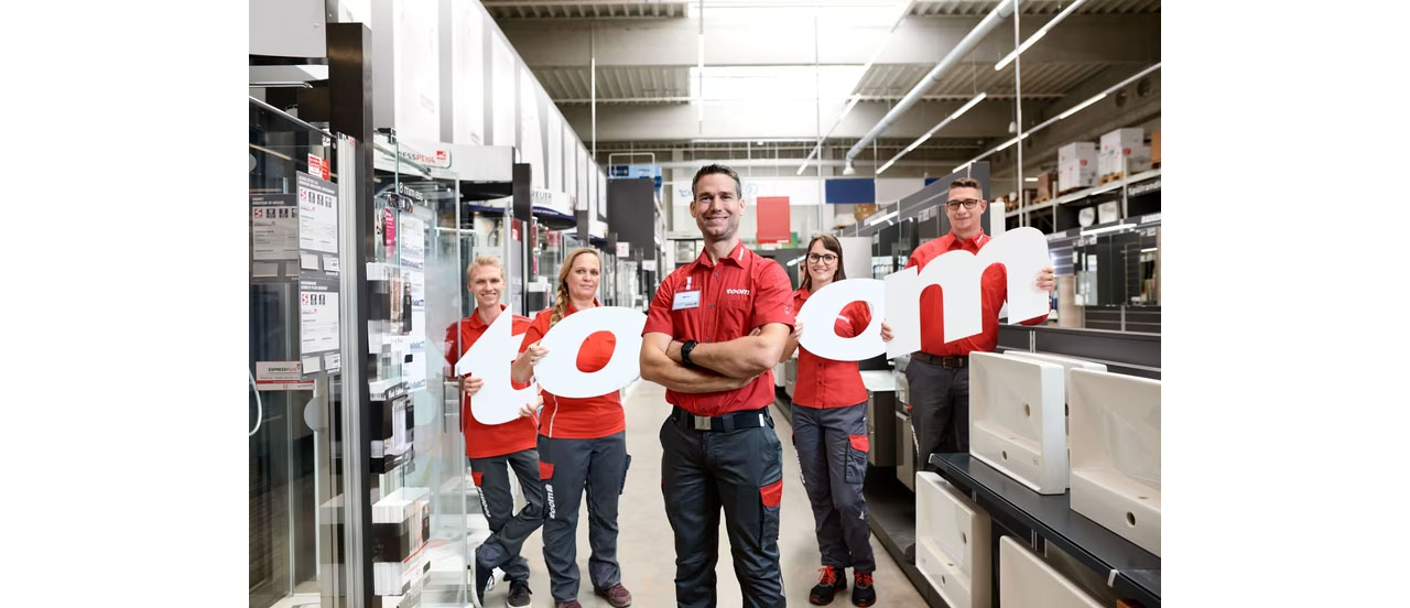 5 Mitarbeitende halten toom-Buchstaben fest und lächeln im Baumarkt stehend in die Kamera