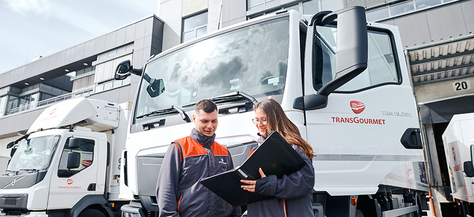Zwei Personen stehen vor einem LKW. Eine Person hält eine Mappe in der Hand, in welche die beiden Personen blicken. Auf der Fahrertür des LKW´s ist das Logo der Firma TRANSGOURMET zu sehen.