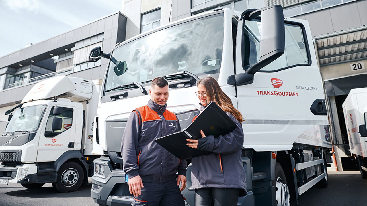 Zwei Personen stehen vor einem LKW. Eine Person hält eine Mappe in der Hand, in welche die beiden Personen blicken. Auf der Fahrertür des LKW´s ist das Logo der Firma TRANSGOURMET zu sehen.