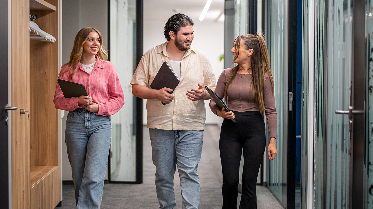 3 junge Mitarbeitende mit laufen mit Unterlagen in den Armen einen Gang in der Steuerkanzlei trilling•hellmann & partner mbB entlang.