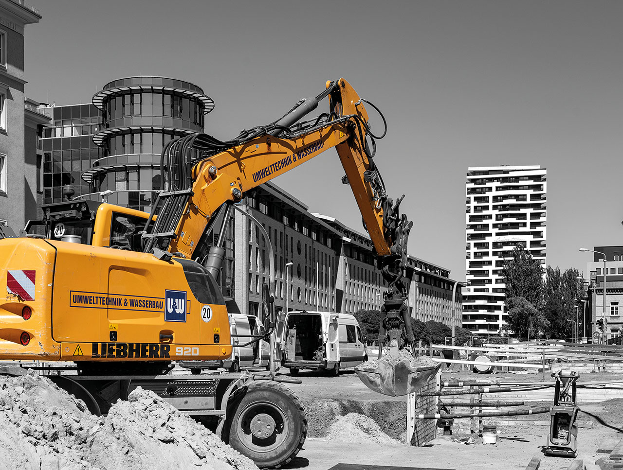 Situation auf einer Baustelle im Vordergrund des Bildes ein großer gelber Bagger, dahinter in schwarz-weiß große Fläche und ein Hochhaus