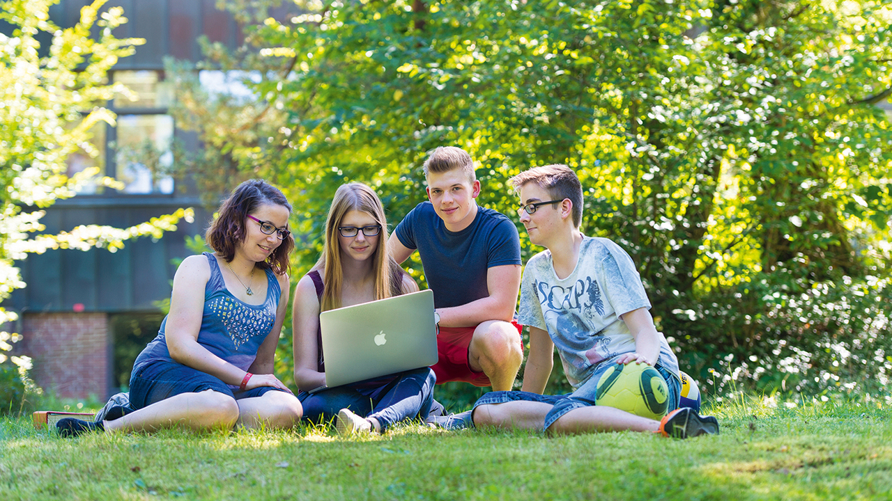 Vier junge Menschen sitzen im Grünen und sehen sich etwas auf dem Laptop an