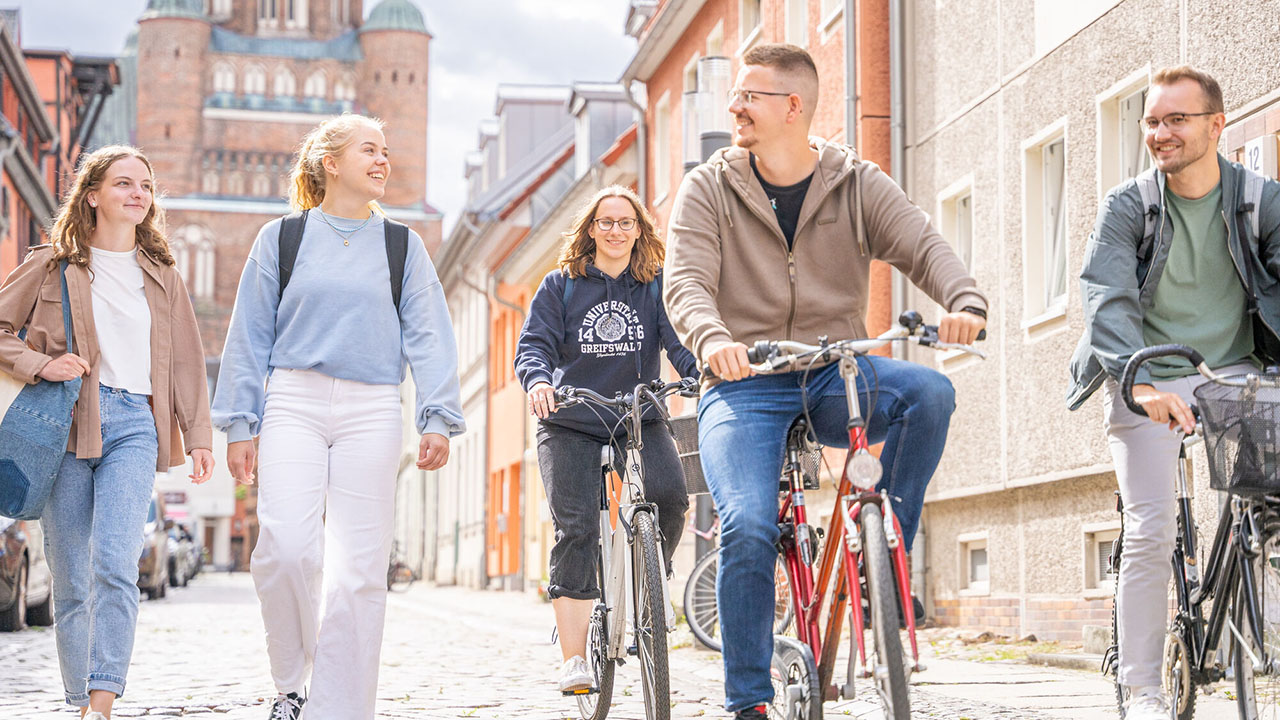 Mehrere junge Personen gehen oder fahren Fahrrad lächelnd durch eine helle Altstadtstraße. Im Hintergrund ist ein großes, altes Gebäude mit Türmen zu sehen.