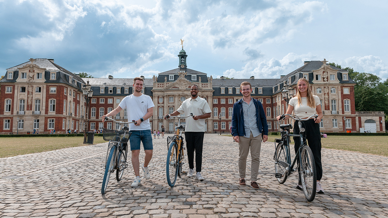 4 Studierende, davon 3 mit Rad vor einem historischen Gebäude der Uni Münster
