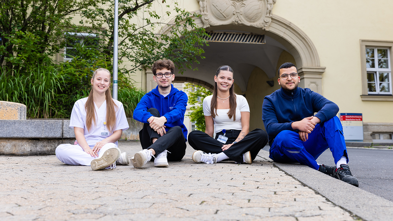 4 junge Mitarbeitende des Universitätsklinikum Würzburg die in Ihrer jeweiligen Arbeitskleidung vor einem Gebäude des Klinikums sitzen und gut gelaunt in die Kamera schauen.