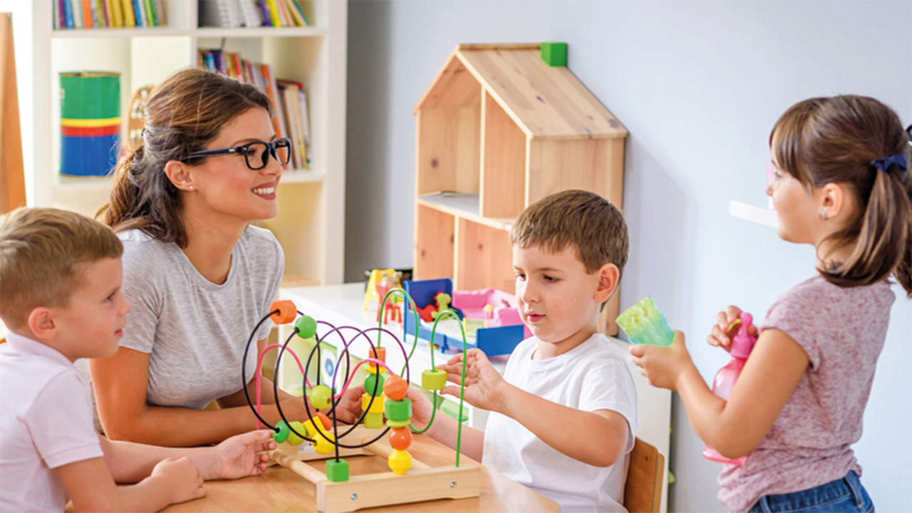 Szene in einer Kita, Erzieherin mit drei Kindern an einem Spieltisch