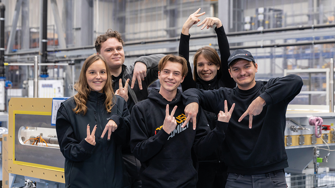 Gruppe von fünf Auszubildenden in einer Werkshalle. Alle schauen fröhlich in die Kamera und formen mit Zeige- und Mittelfinger ein V für von und ein A für Ardenne