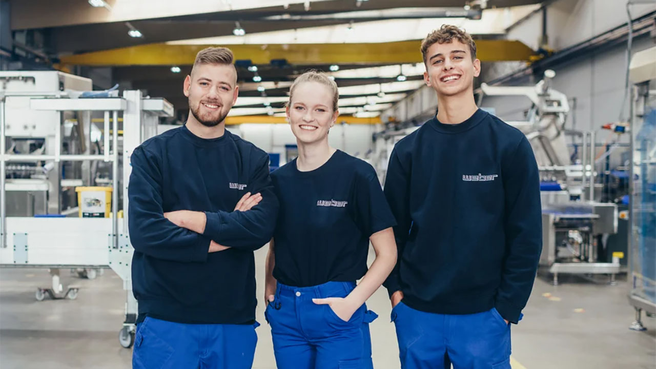 Das Bild zeigt drei Personen die in die Kamera lächeln. Im Hintergrund sieht man eine Werkshalle.