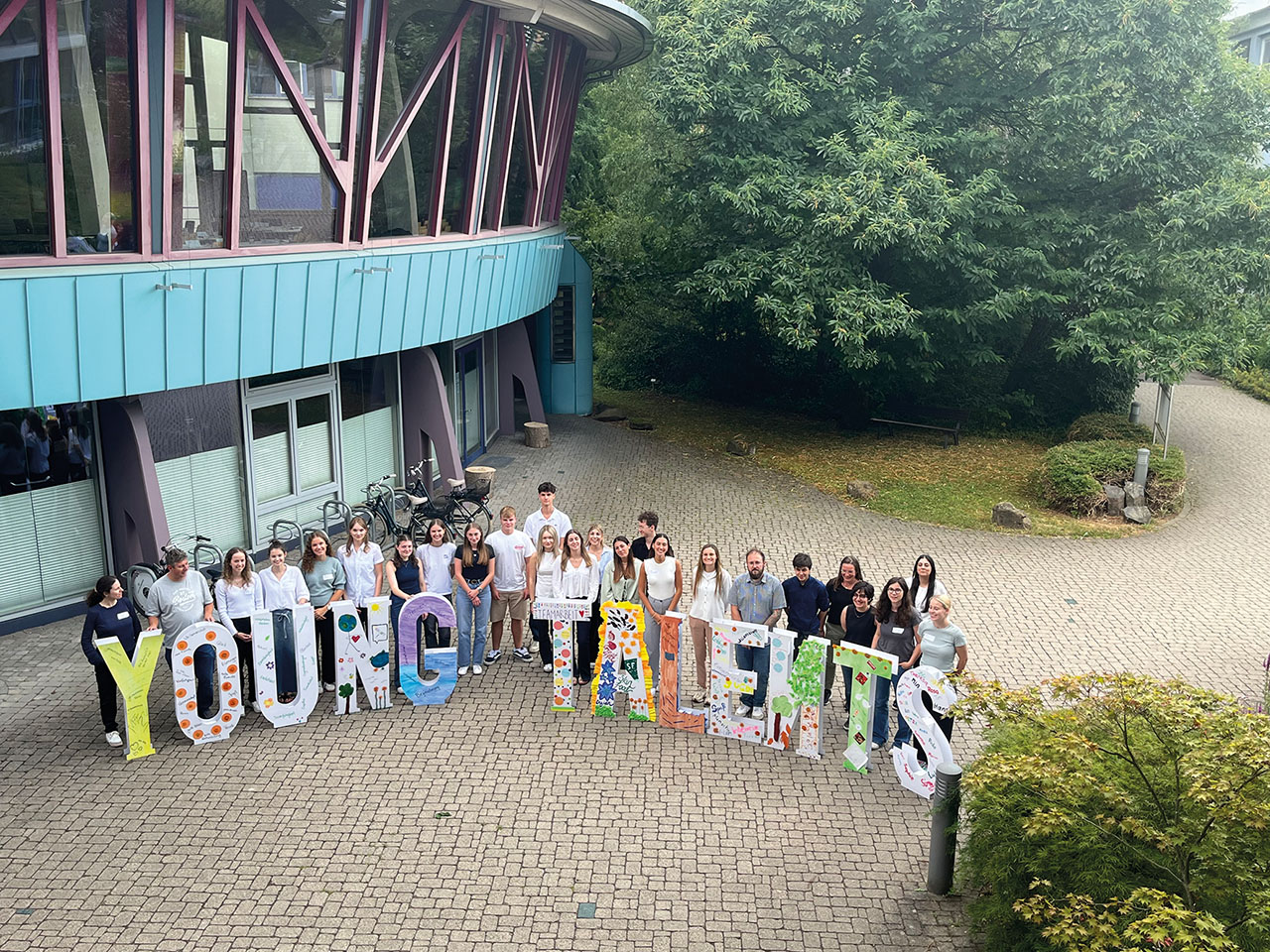 Eine Gruppe von Personen steht draußen vor einem Gebäude. Vor den Personen stehen große bunte Buchstaben, die zusammen „YOUNG TALENTS“ ergeben.