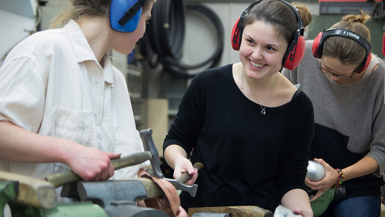 Drei junge Menschen in einer Werkstatt beim Bearbeiten von Metall mit einem Hammer.
