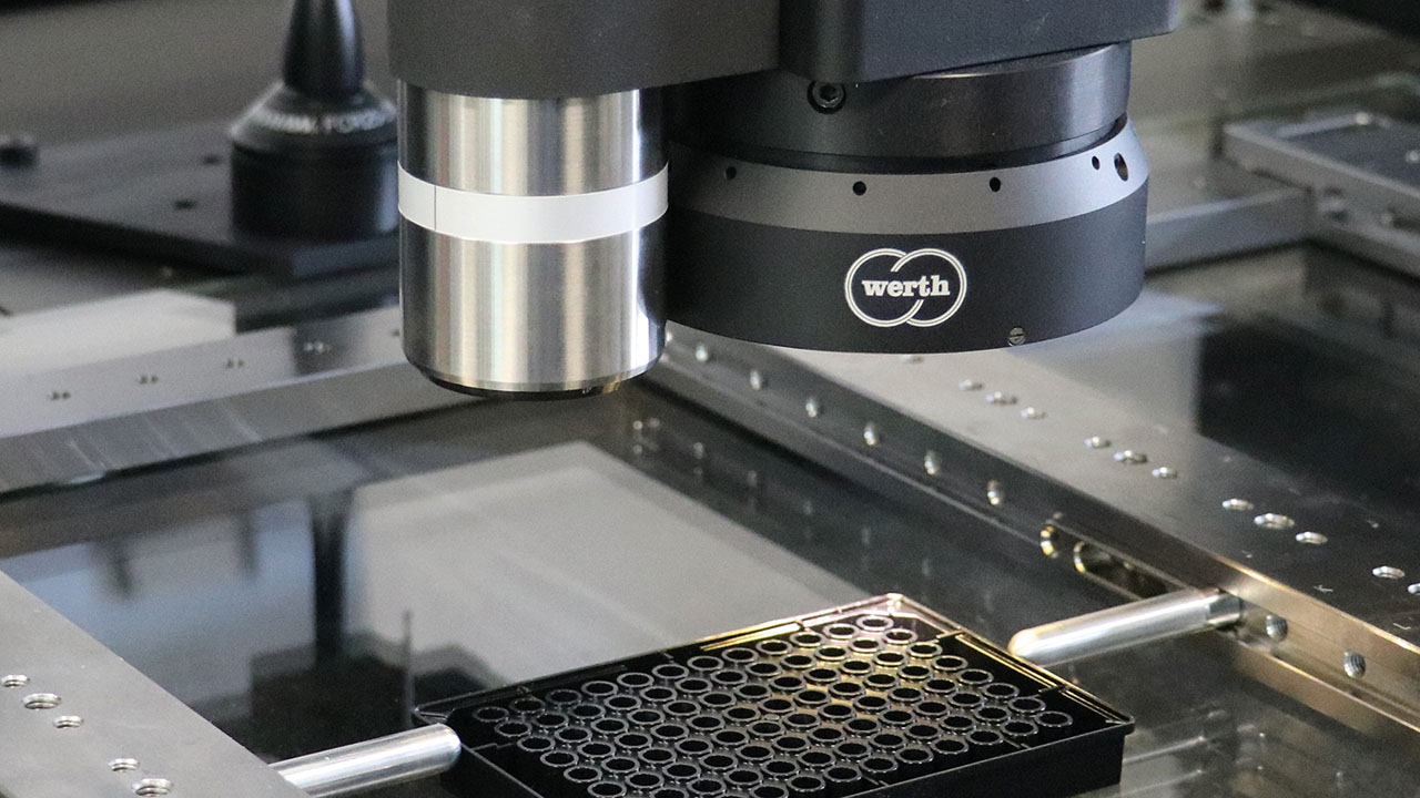 Das Bild zeigt einen industriellen Mess- oder Prüfkopf der Marke Werth, der über einer Metallplattform positioniert ist. Darunter befindet sich eine schwarze Halterung mit vielen kleinen zylindrischen Bauteilen – vermutlich zur präzisen Vermessung oder Qualitätskontrolle in der Fertigung.