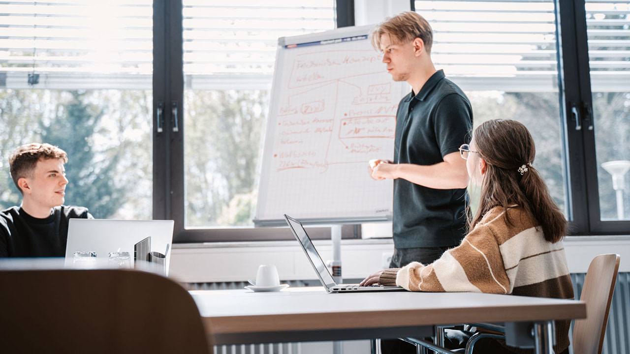 Drei Personen am Schreibtisch bzw. vor einem Flipchart. 