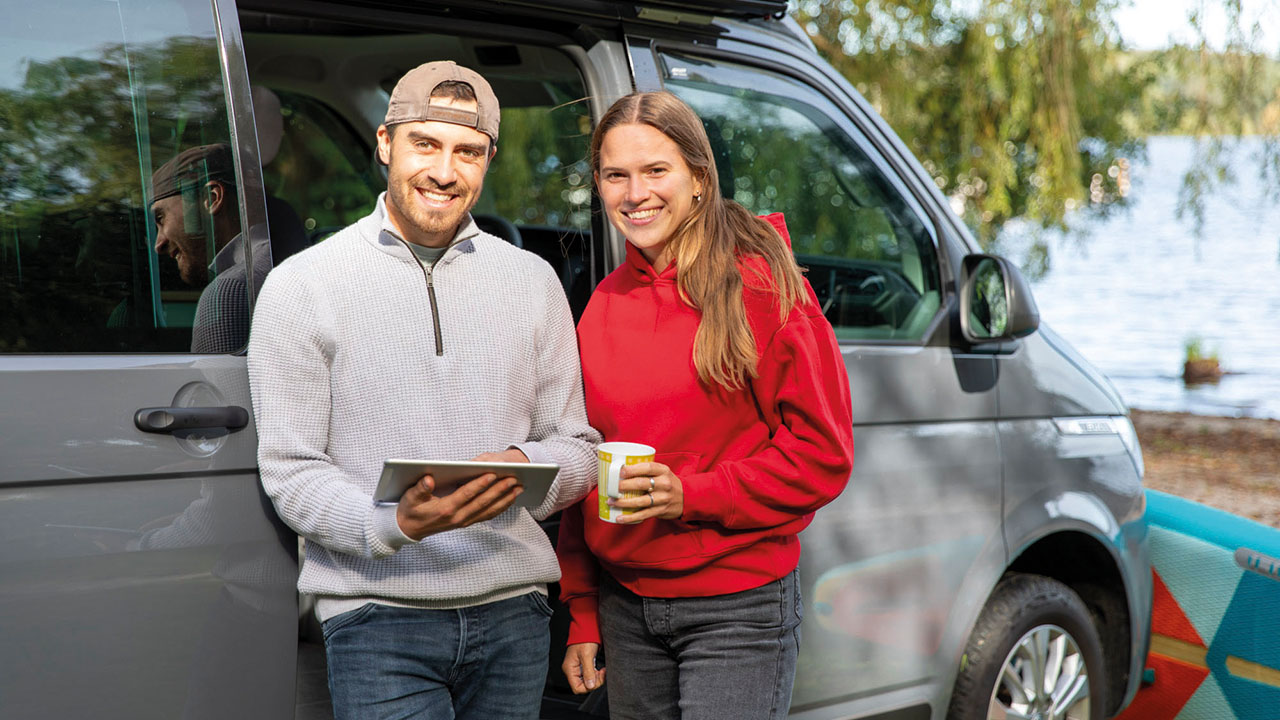 Zwei junge Menschen stehen vor Kleinbus mit SUP im Hintergrund am Strand und lächeln in die Kamer.