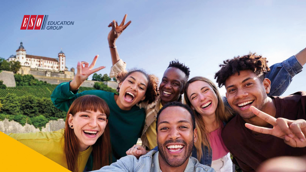 Gruppenfoto von fünf jungen Erwachsenen, die fröhlich in die Kamera lachen und Peace-Zeichen machen. Im Hintergrund ist eine historische Burg inmitten von Weinbergen zu sehen. Links oben steht das Logo „ESO Education Group“.