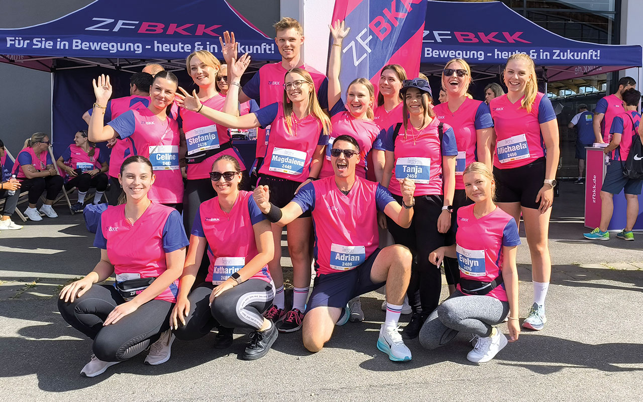 Eine Gruppe von Personen trägt sportliche Kleidung in Pink und Blau und posiert draußen vor einem Zelt mit der Aufschrift "ZF BKK". Einige stehen, andere sitzen in der Hocke oder knien. Alle tragen Startnummern.